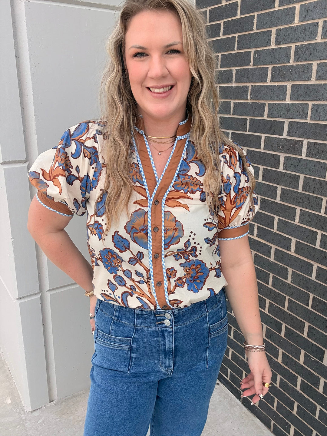 A woman wearing a short-sleeve button-up blouse with a blue and brown floral pattern on a cream background, paired with blue jeans, standing outdoors against a gray brick wall.