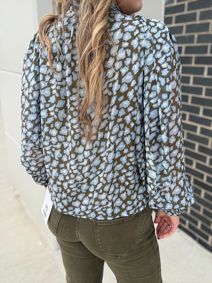 A woman shown from the back wearing a light blue and olive green leopard-print blouse with long sleeves, paired with olive green pants, standing outdoors next to a brick wall.