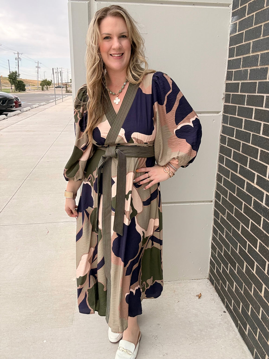 A woman wearing a long-sleeve maxi dress with a bold abstract floral pattern in navy blue, olive green, beige, and pink, standing outdoors on a concrete sidewalk next to a gray wall and brick column.