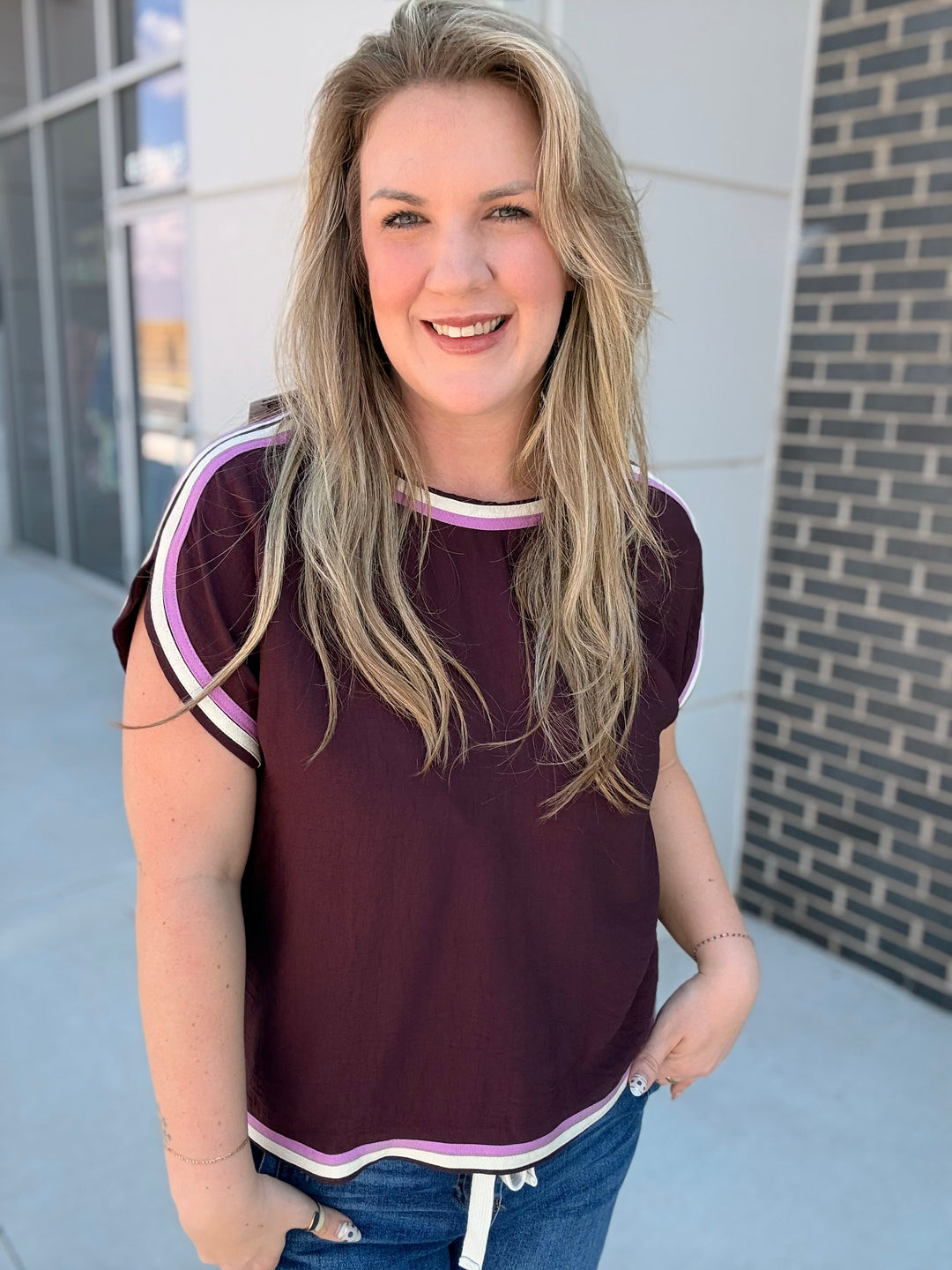 A woman with blonde hair wearing a short-sleeve crewneck top in deep burgundy with white and light purple striped trim, paired with blue jeans, standing outdoors in front of a modern building.