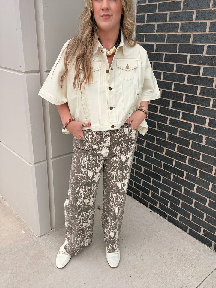A woman stands outdoors against a gray concrete wall and dark brick wall, wearing a cream short-sleeve button-up shirt with brown buttons and patterned gray-and-white pants, paired with white pointed shoes.