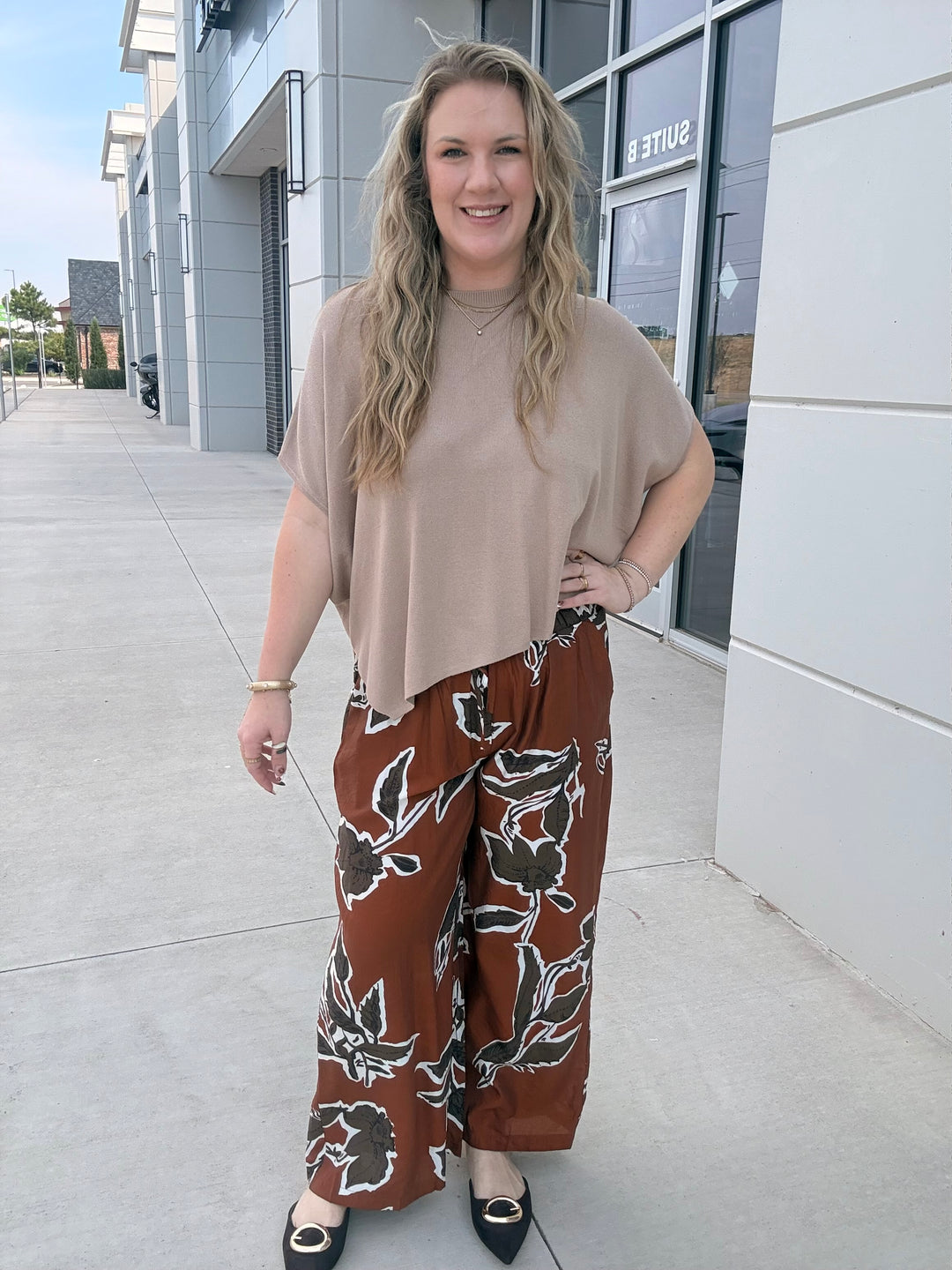 A woman stands outdoors on a sidewalk in front of a modern building, wearing a loose beige short-sleeve top and wide-leg brown pants with a black and white floral pattern.