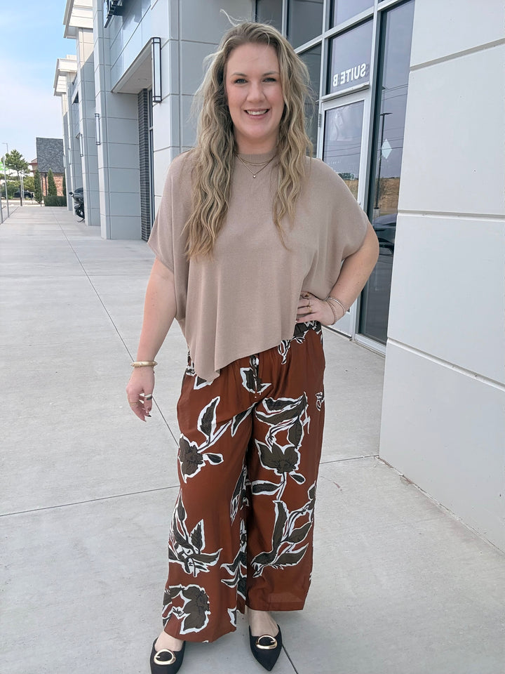 A woman stands outdoors on a sidewalk in front of a modern building, wearing a loose beige short-sleeve top and wide-leg brown pants with a black and white floral pattern.