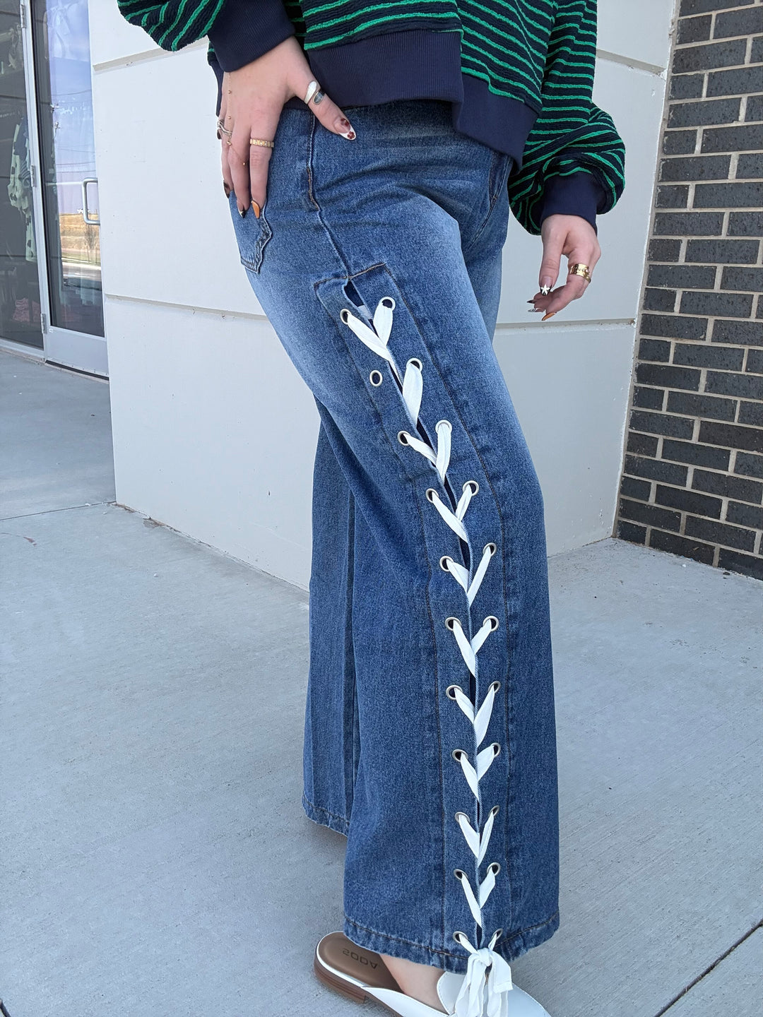 A person wearing blue wide-leg jeans with white lace-up detailing down the front of each leg, paired with a green and navy striped sweater and white slip-on shoes, standing outdoors on a sidewalk.