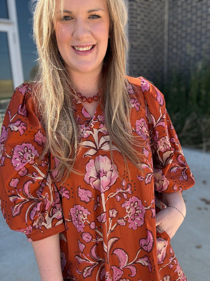 A woman wearing a rust-orange short-sleeve blouse with a pink and beige floral pattern, standing outdoors.
