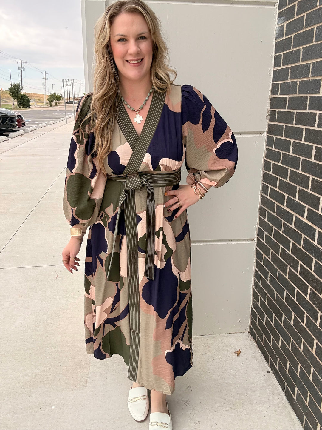 A woman modeling a long-sleeve wrap maxi dress with a camouflage pattern in navy blue, olive green, and beige, standing outdoors next to a gray wall and brick column.