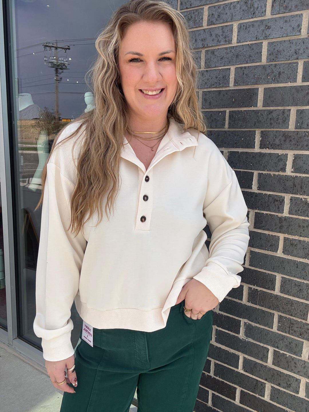 A woman wearing a cream-colored long-sleeve henley sweatshirt and dark green pants, standing outdoors against a dark gray brick wall.