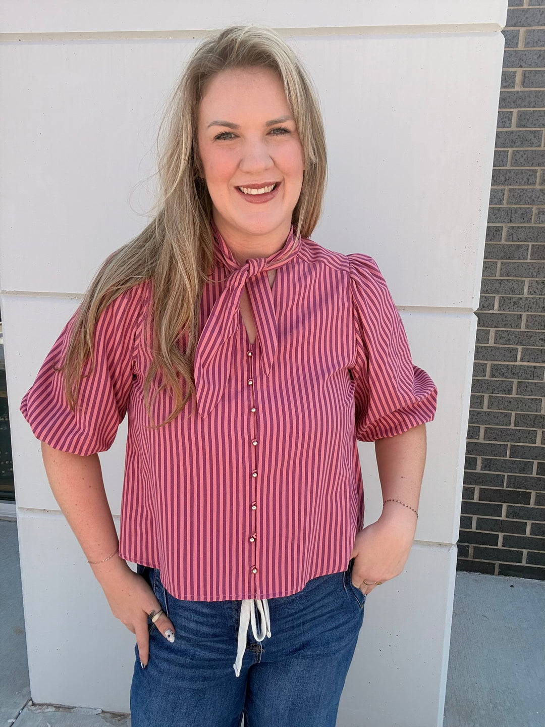 A smiling blonde woman wearing a pink and red vertically striped short-sleeve blouse with puff sleeves and a tie-neck detail, paired with blue jeans, standing outdoors against a white wall.