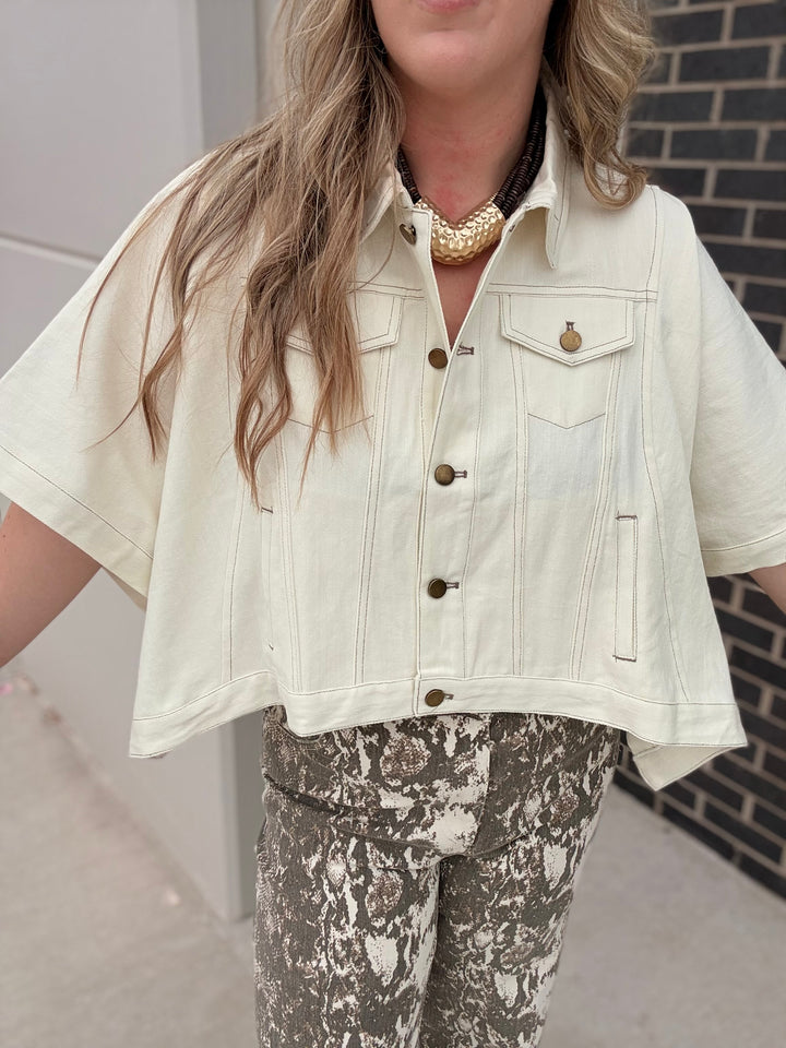 A woman wearing a cream-colored, cropped denim jacket with brown buttons and a small gold chest emblem, paired with brown and white patterned pants.