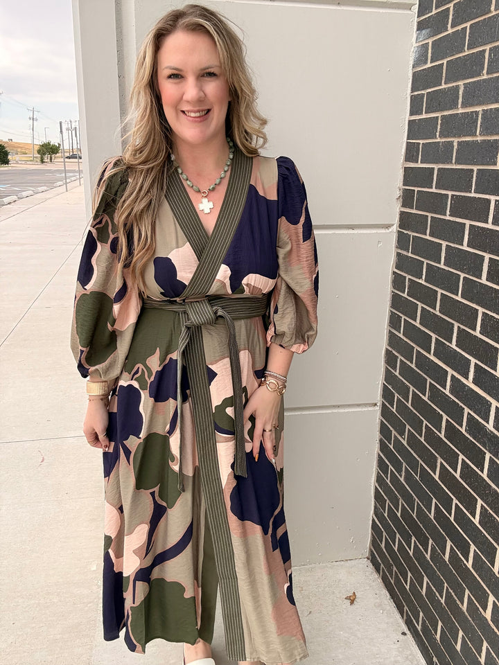 A woman wearing a long-sleeve wrap maxi dress with a camouflage pattern in olive green, beige, and navy blue, standing outdoors next to a brick wall.