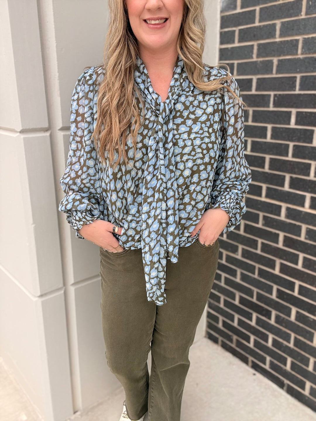 A woman wearing a light blue and olive green leopard print blouse with long sleeves and a tie-neck detail, paired with olive green pants, standing outdoors against a white wall and a dark brick wall.