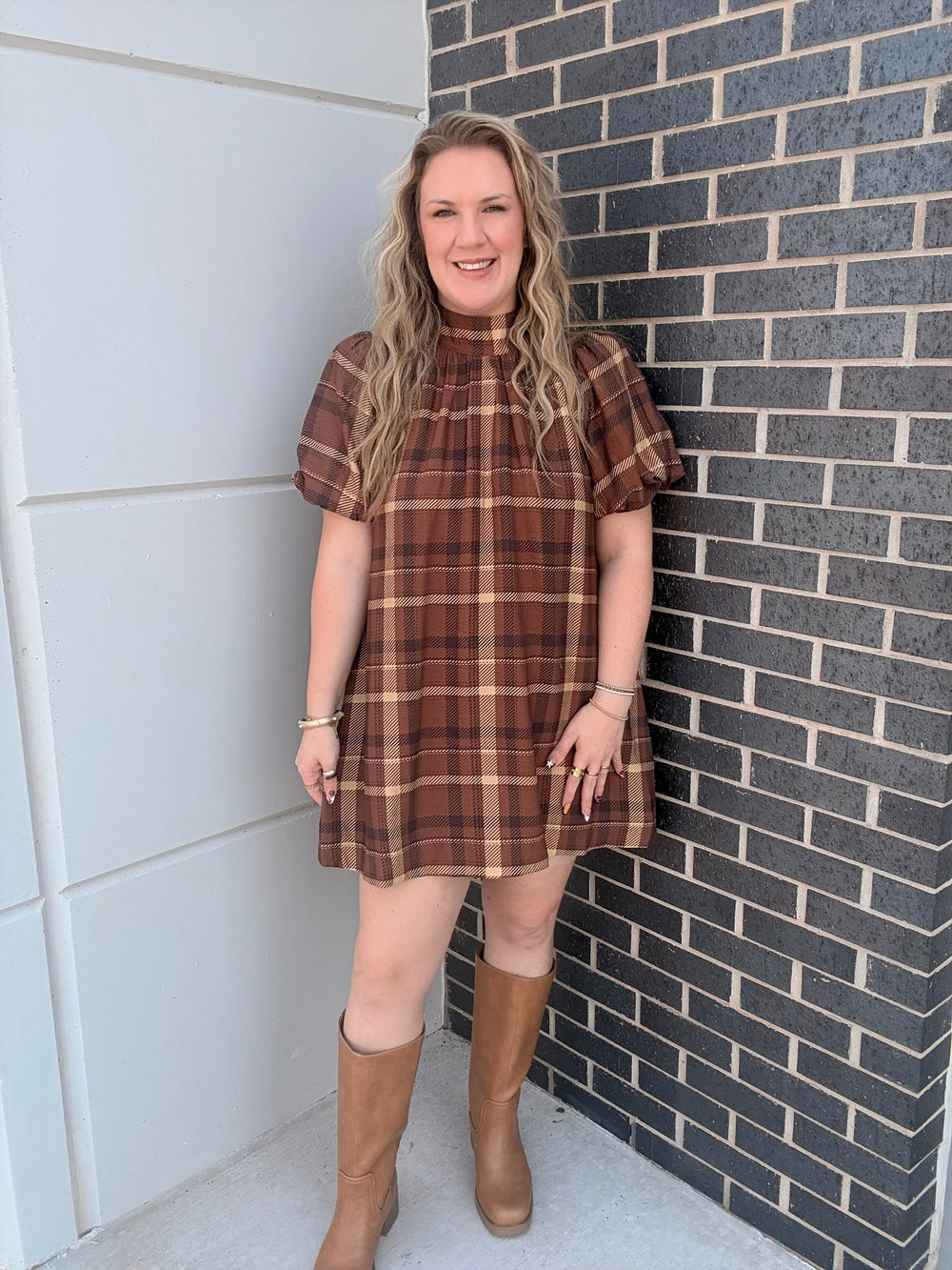 A woman wearing a brown and tan plaid short-sleeve mini dress with puff sleeves, standing outdoors against a gray brick wall and white paneled wall.