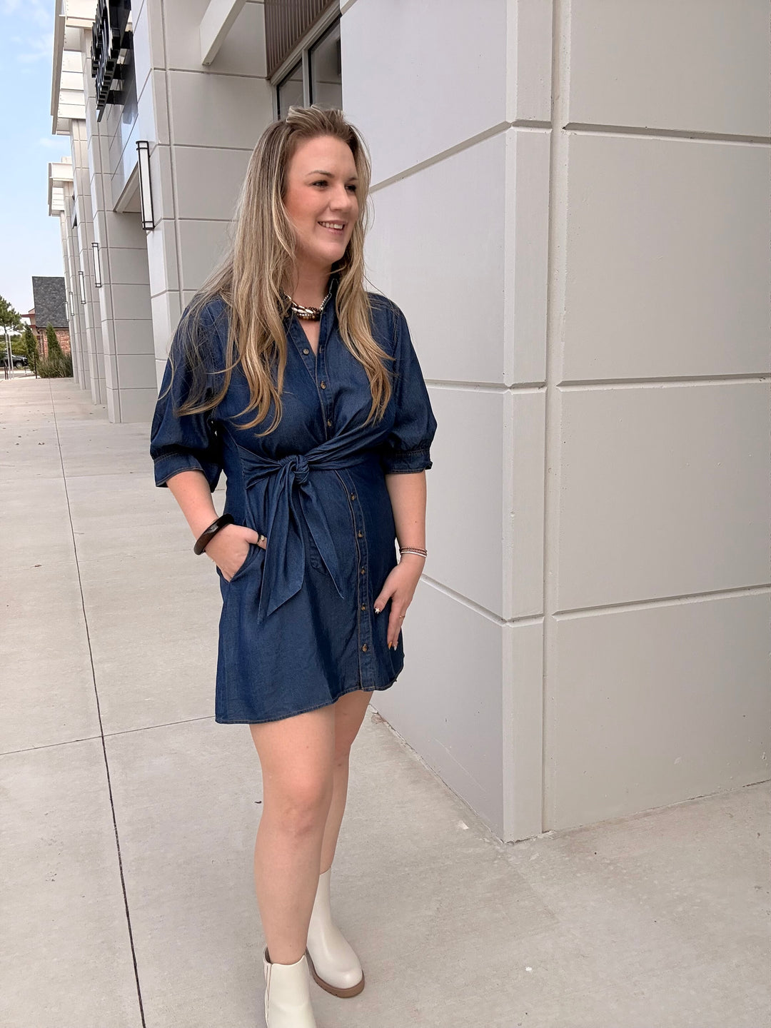 A woman wearing a dark blue denim mini dress with a tie waist and white ankle boots, standing outdoors next to a modern light gray building.