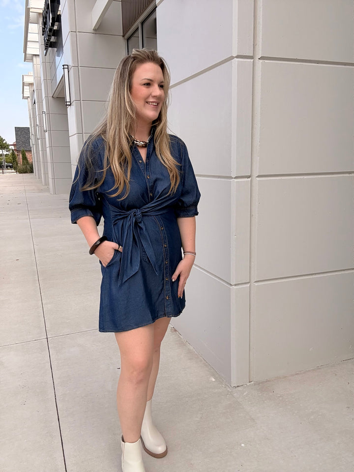 A woman wearing a dark blue denim mini dress with a tie waist and white ankle boots, standing outdoors next to a modern light gray building.