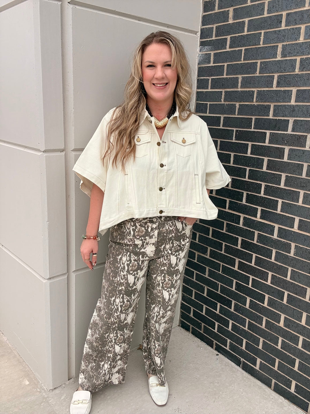 A woman stands outdoors wearing a cream short-sleeve button-up jean shirt and gray-and-white snakeskin print pants with white sneakers.