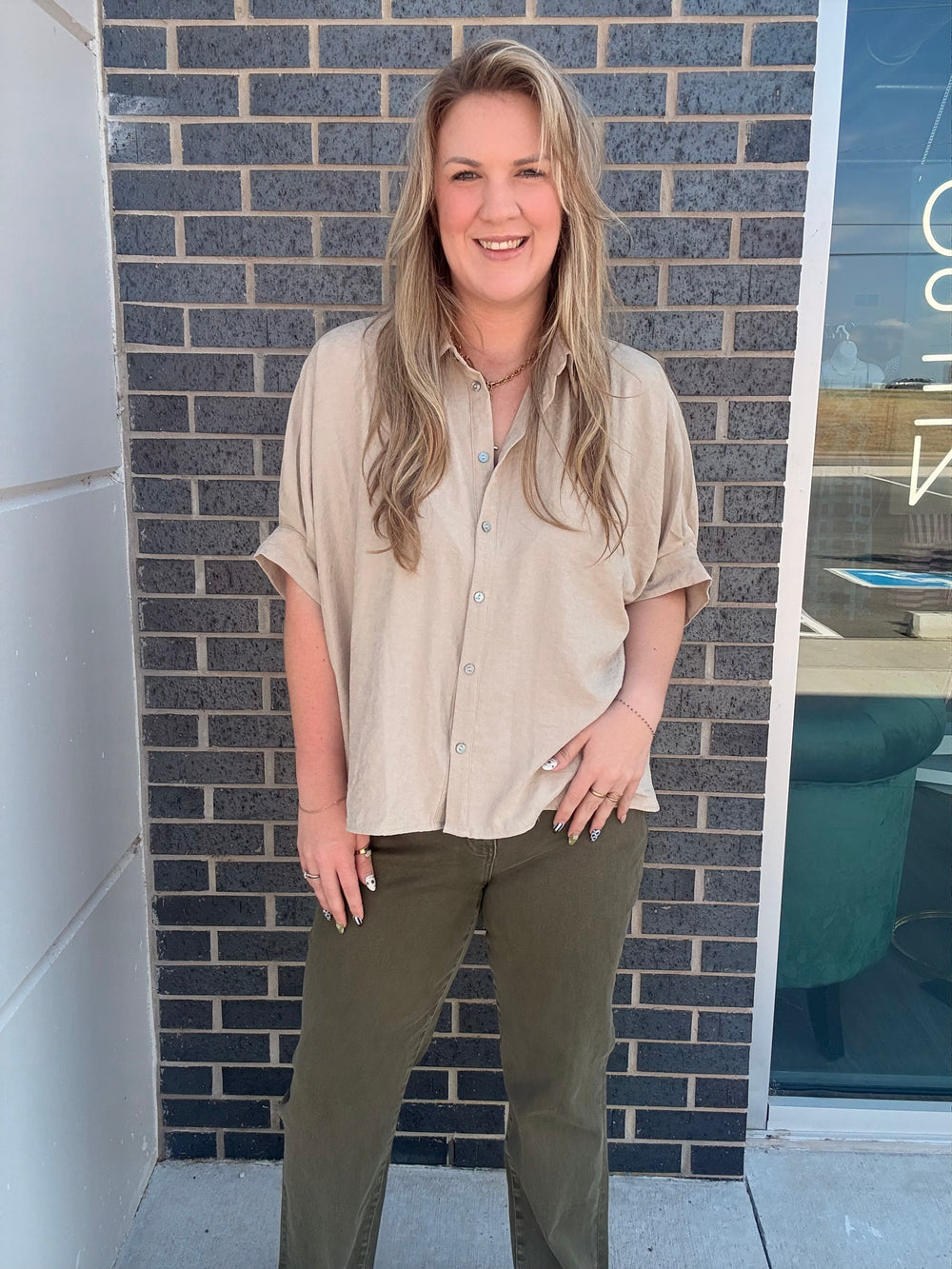 A woman with blonde hair stands in front of a dark gray brick wall, wearing a light beige short-sleeve button-up shirt and olive green pants.