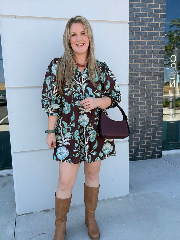 A woman stands outdoors wearing a brown mini dress with a teal and white floral pattern, paired with tan knee-high boots and a dark plum shoulder bag.