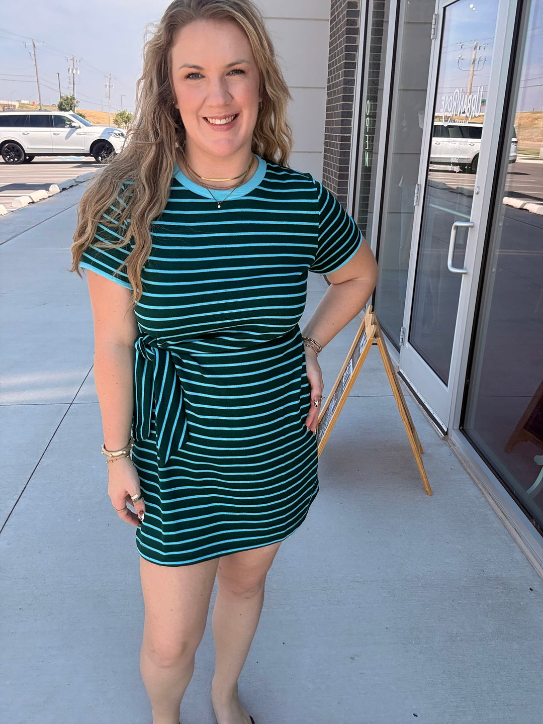 A woman modeling a short-sleeve mini dress with dark green and white horizontal stripes, featuring a light blue collar and a matching light blue tie waist, standing outdoors on a sidewalk.