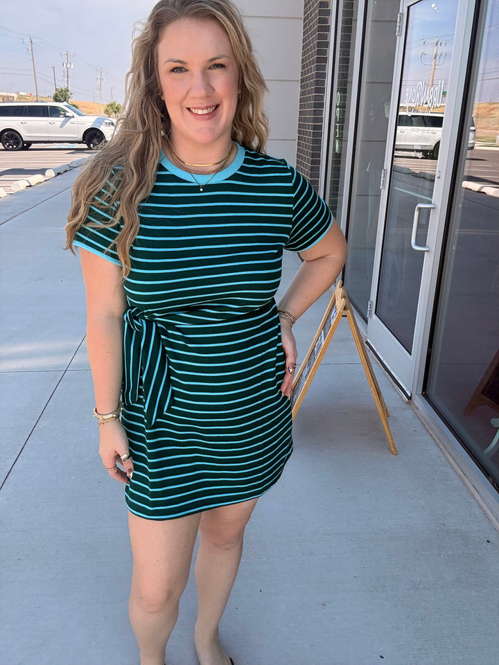 A woman modeling a short-sleeve mini dress with dark green and white horizontal stripes, featuring a light blue collar and a matching light blue tie waist, standing outdoors on a sidewalk.