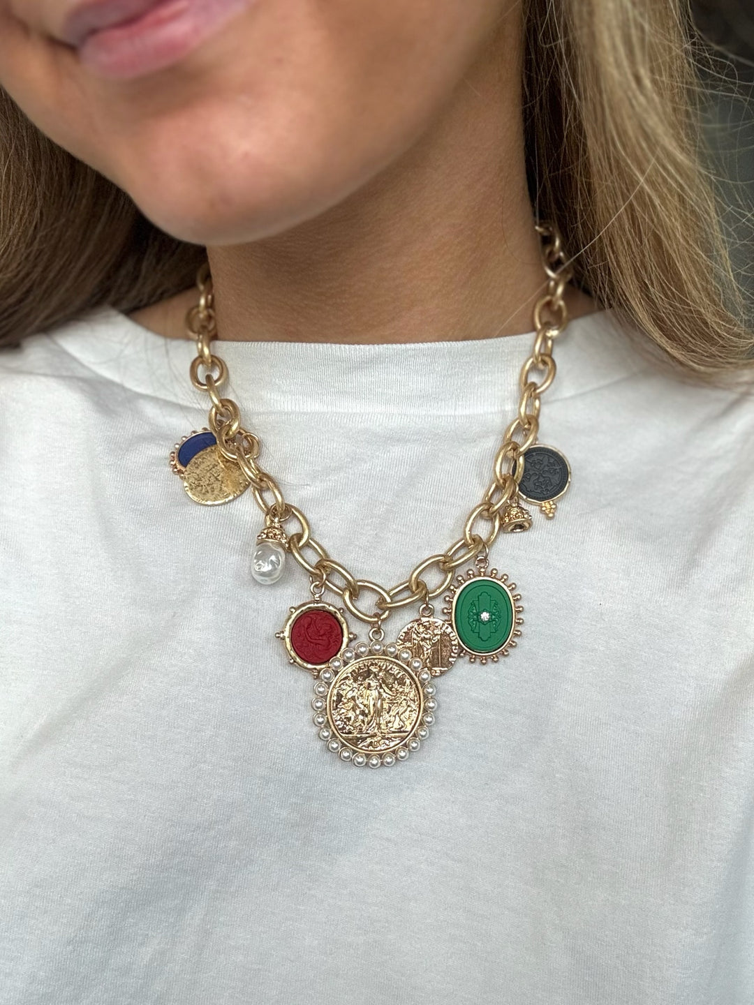 A close-up of a person wearing a gold-tone charm necklace with multiple oval and round pendants in red, green, blue, and dark gray, styled over a white t-shirt.
