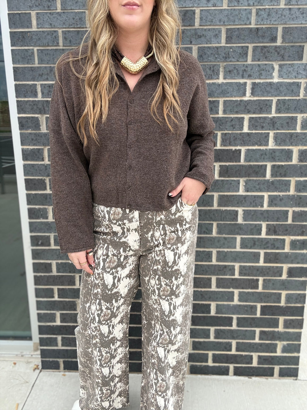 A woman wearing a brown fuzzy long-sleeve top and brown-and-white patterned wide-leg pants, standing in front of a gray brick wall.