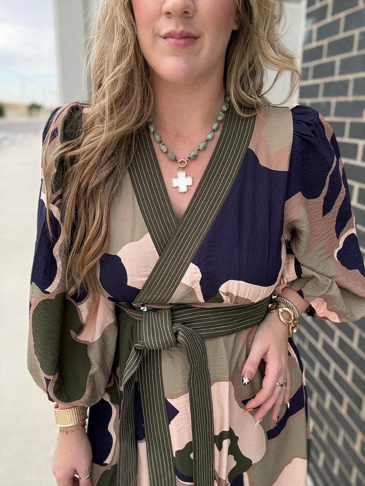 A woman wearing a wrap-style dress with a camouflage pattern in navy, olive green, beige, and brown, featuring a matching wide waist tie belt. She is wearing a green beaded necklace with a white cross pendant.