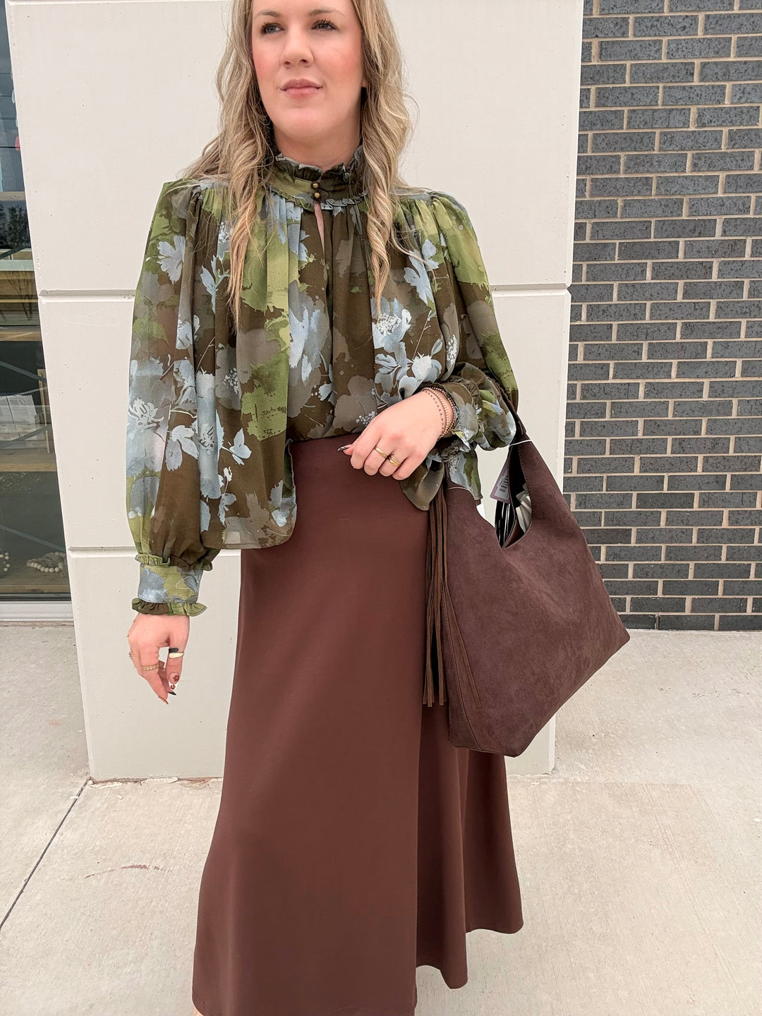 A woman wearing a green and brown floral long-sleeve blouse with a high neck, paired with a brown midi skirt and carrying a large brown tote bag, standing outdoors against a white wall and brick column.