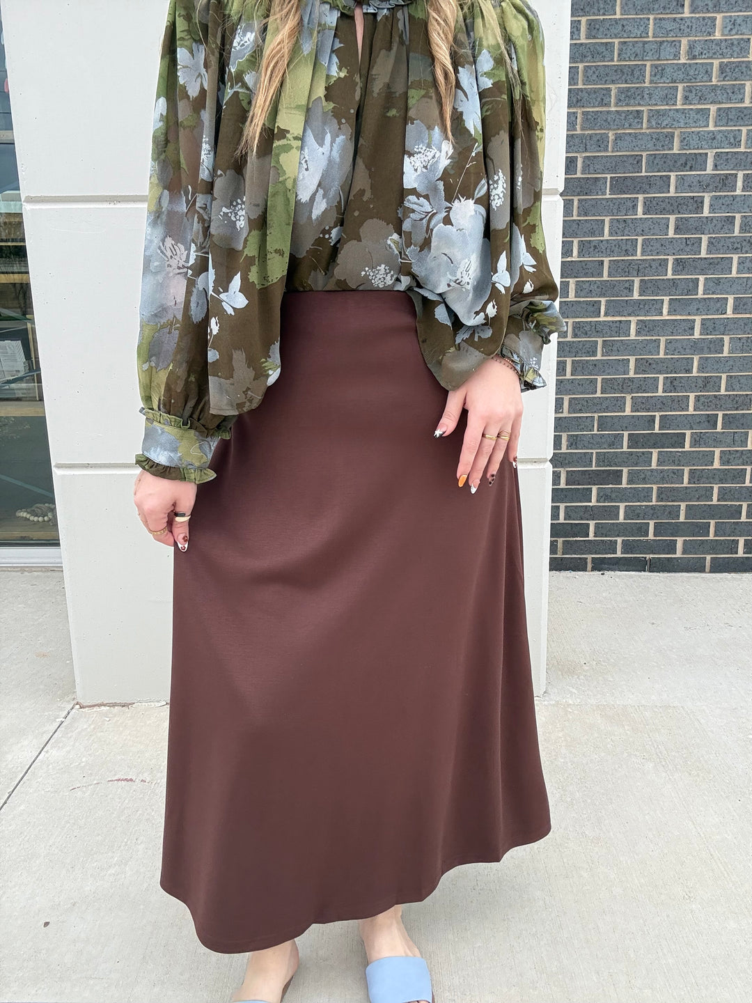A person wearing a brown midi skirt and a green floral blouse, standing outdoors against a white wall and dark brick wall.