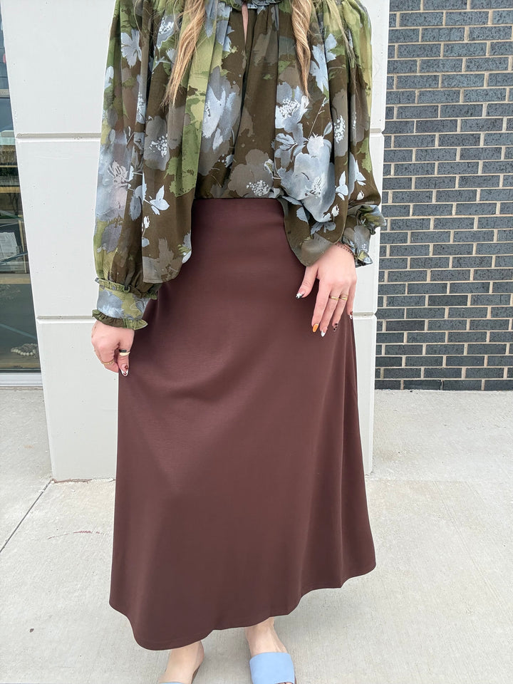 A person wearing a brown midi skirt and a green floral blouse, standing outdoors against a white wall and dark brick wall.