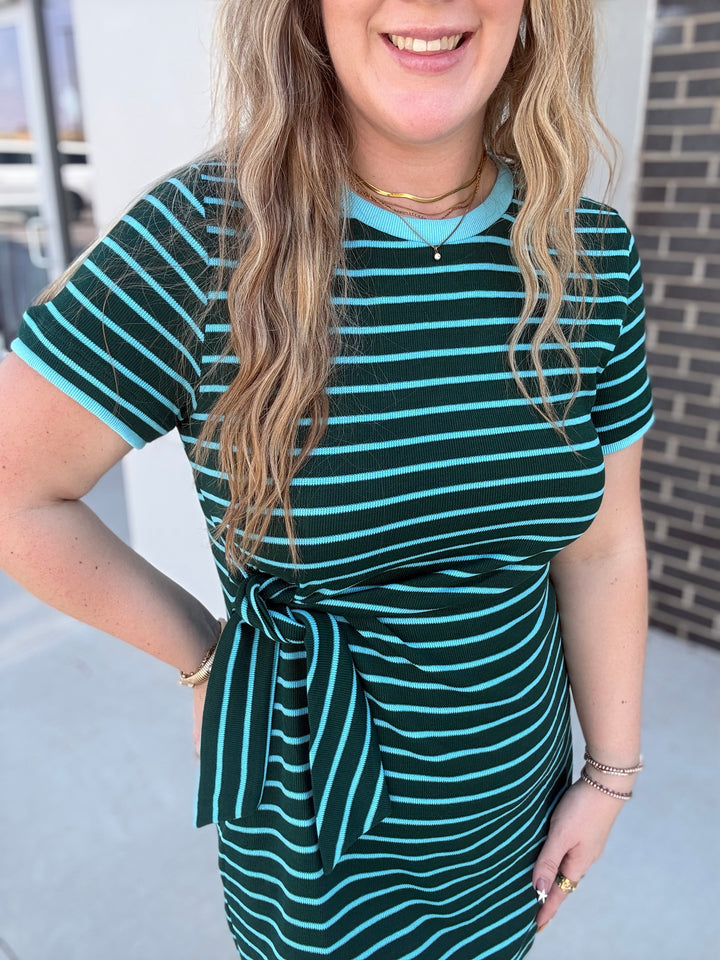 A woman wearing a short-sleeve knit dress with horizontal teal and dark green stripes, featuring a tie-waist detail and a crew neckline.