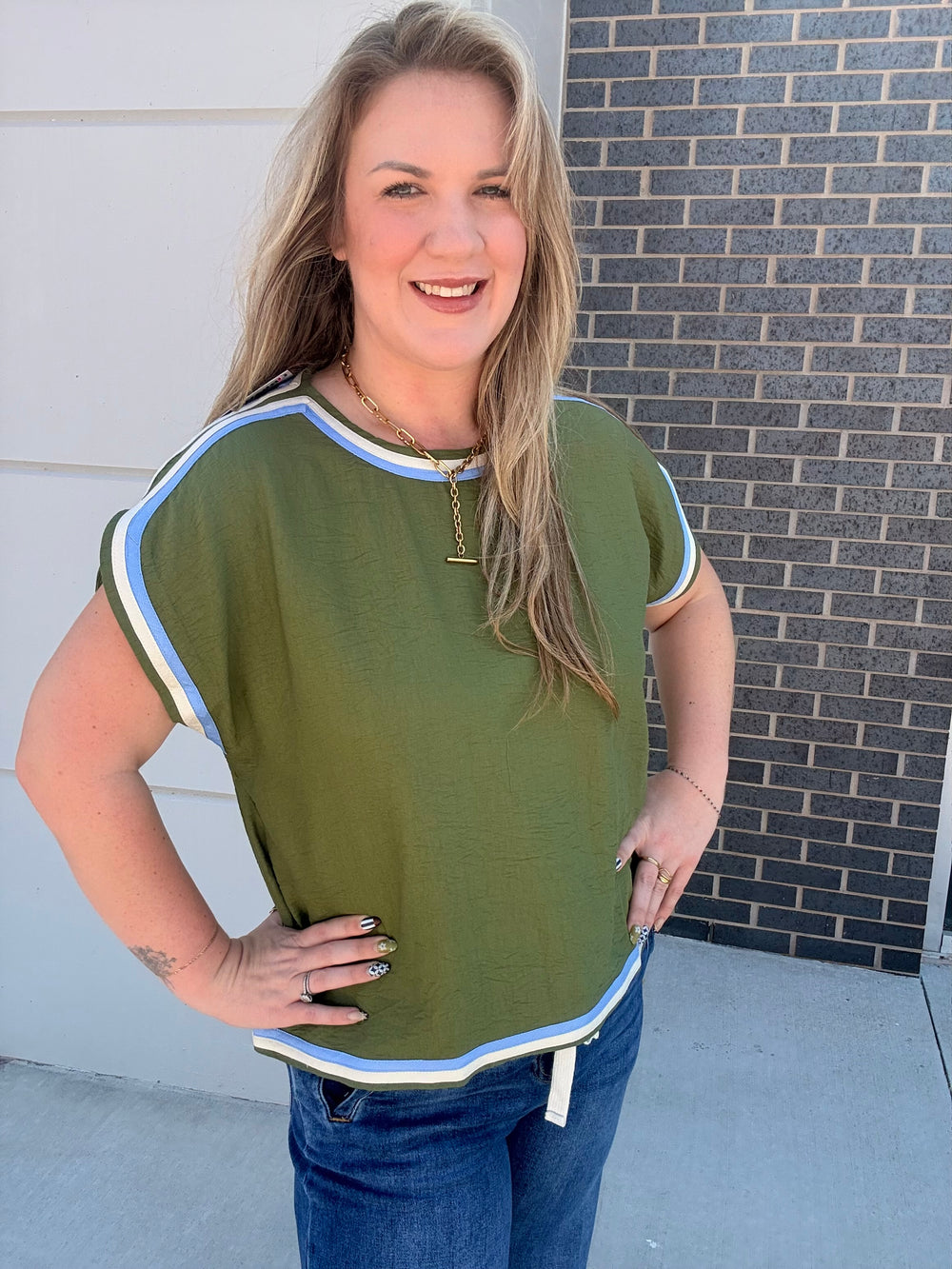 A woman wearing an olive green short-sleeve top with light blue and white striped trim, paired with blue jeans, standing outdoors against a white wall and a dark brick wall.