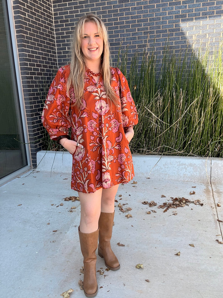 A woman stands outdoors wearing a loose, short-sleeve orange-red floral dress with pink and white flowers, paired with tan knee-high boots. The background shows a gray brick wall, tall green ornamental grass, and a concrete patio.
