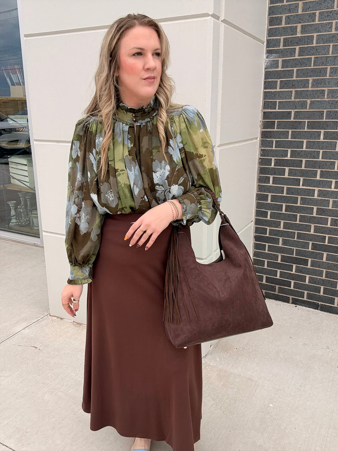 A woman stands outdoors wearing a green and brown floral long-sleeve blouse with a black ruffled collar, a brown midi skirt, and carrying a large brown suede-like shoulder bag.