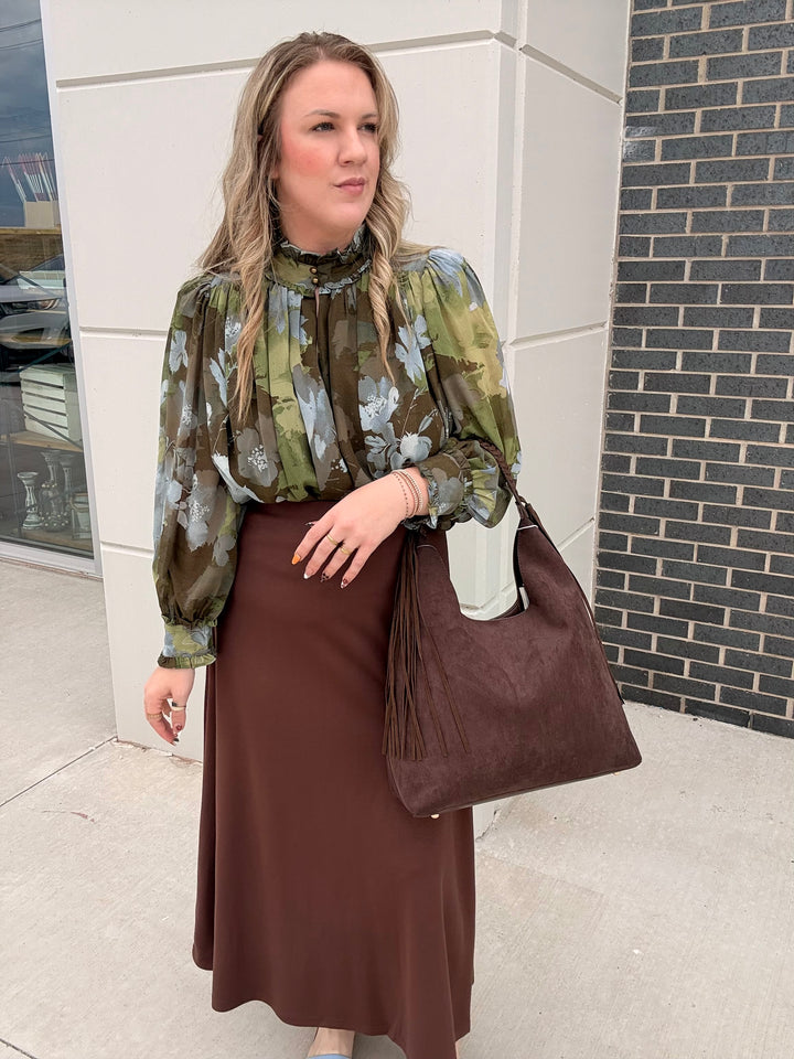 A woman stands outdoors wearing a green and brown floral long-sleeve blouse with a black ruffled collar, a brown midi skirt, and carrying a large brown suede-like shoulder bag.