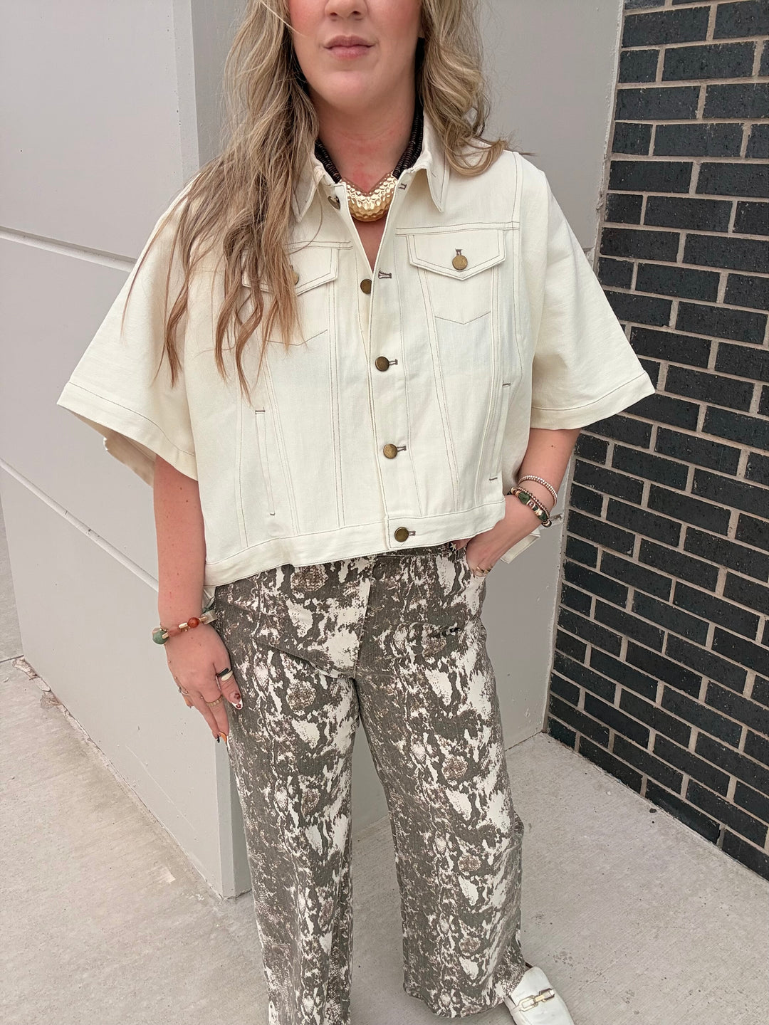 A woman wearing a cream short-sleeve cropped denim jacket and gray-and-white patterned wide-leg pants, standing outdoors next to a white wall and a black brick wall.