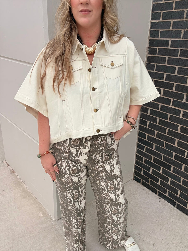A woman wearing a cream short-sleeve cropped denim jacket and gray-and-white patterned wide-leg pants, standing outdoors next to a white wall and a black brick wall.