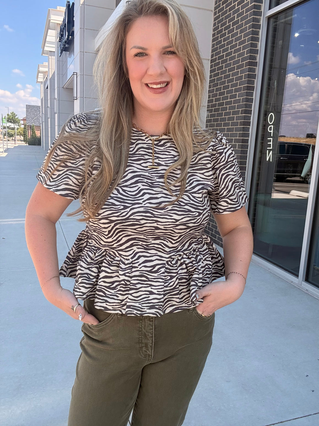 A woman wearing a short-sleeve peplum top with a black and white zebra print, paired with olive green pants, standing outdoors in front of a commercial building.