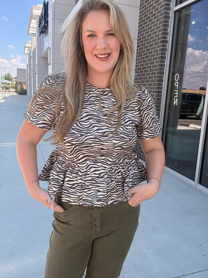 A woman wearing a short-sleeve peplum top with a black and white zebra print, paired with olive green pants, standing outdoors in front of a commercial building.