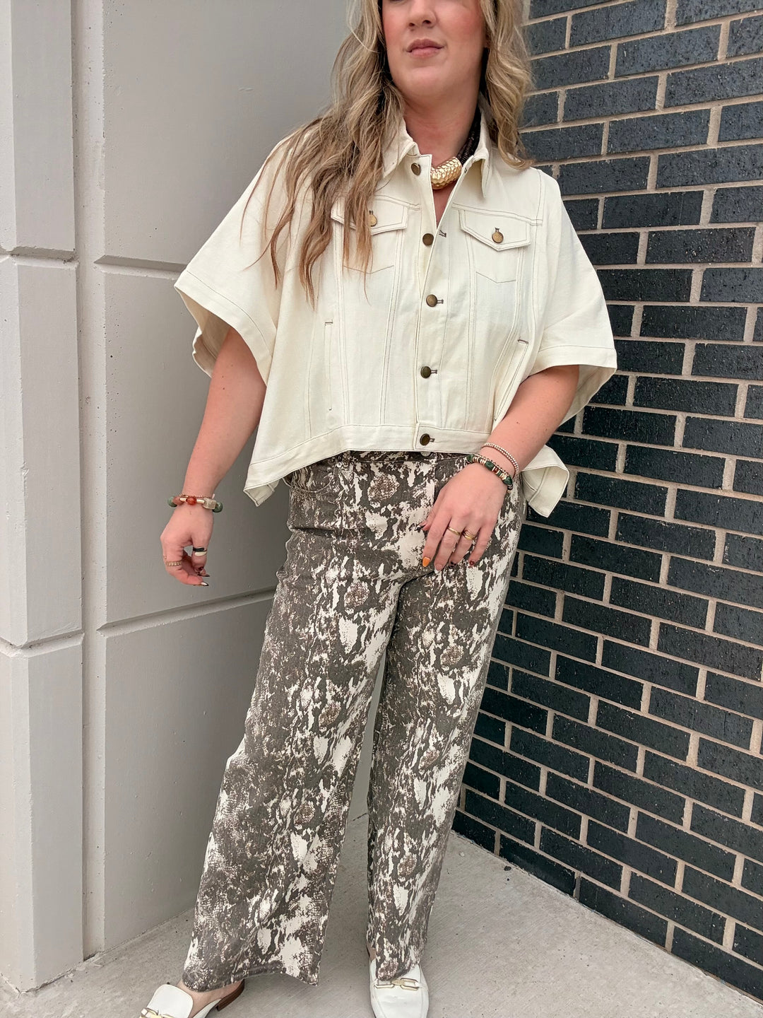 A woman wearing a cream short-sleeve button-up shirt and gray and white snakeskin wide-leg pants, standing outdoors against a gray paneled wall and a dark brick wall.