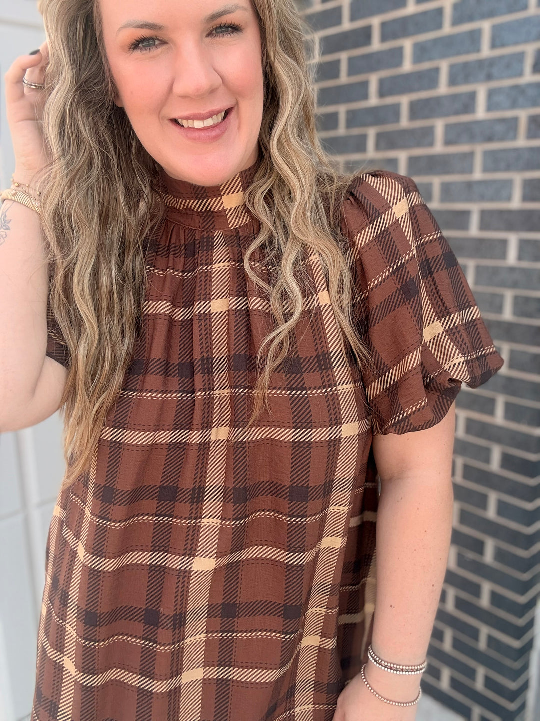 A woman wearing a brown and beige plaid short-sleeve top, standing in front of a gray brick wall.