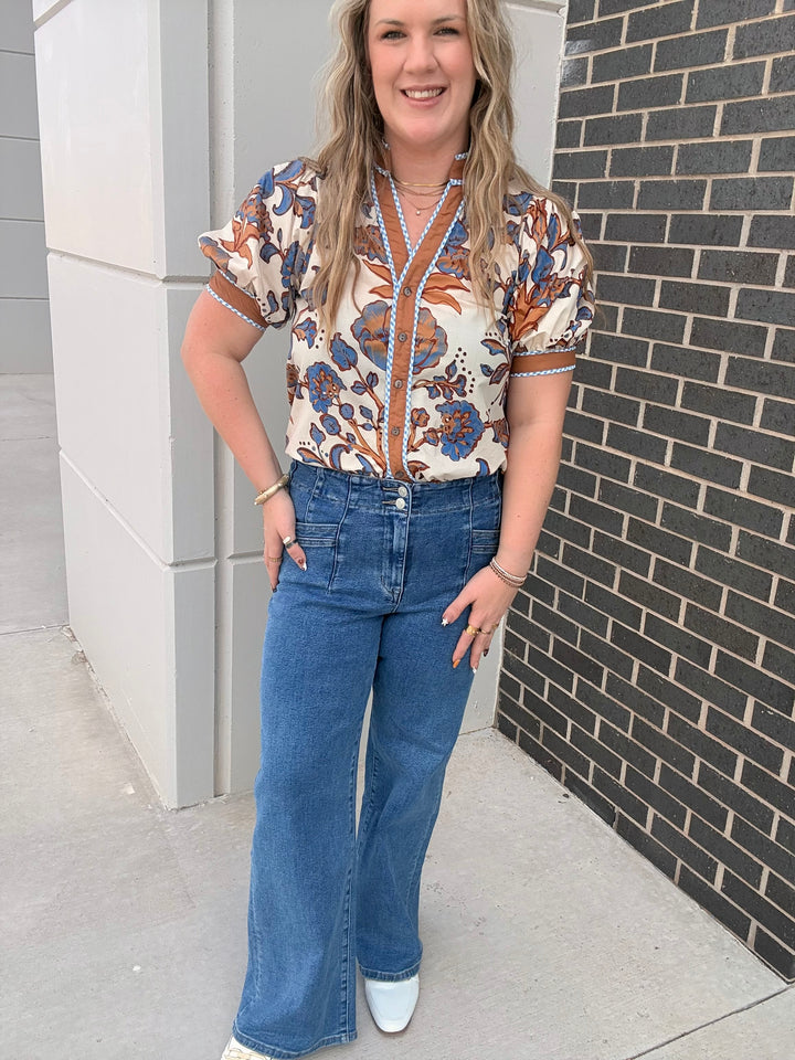 A woman modeling a short-sleeve button-front blouse with a cream base and orange, blue, and brown floral pattern, paired with high-waisted blue flare jeans and white shoes, standing outdoors against a gray concrete wall and dark brick wall.