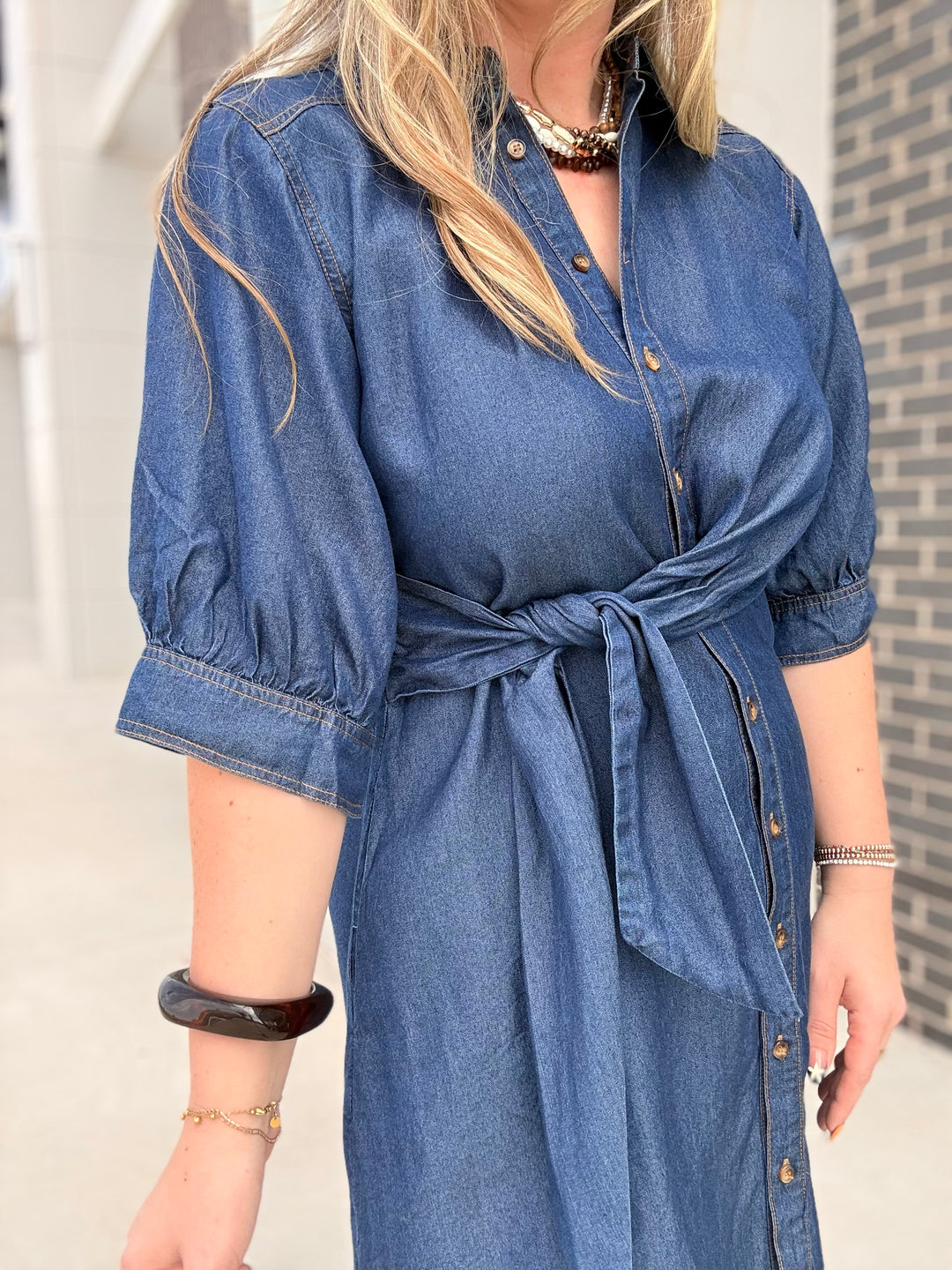 A woman wearing a blue denim shirt dress with puff sleeves, a button front, and a tie waist, photographed outdoors against a light wall and brick wall background.