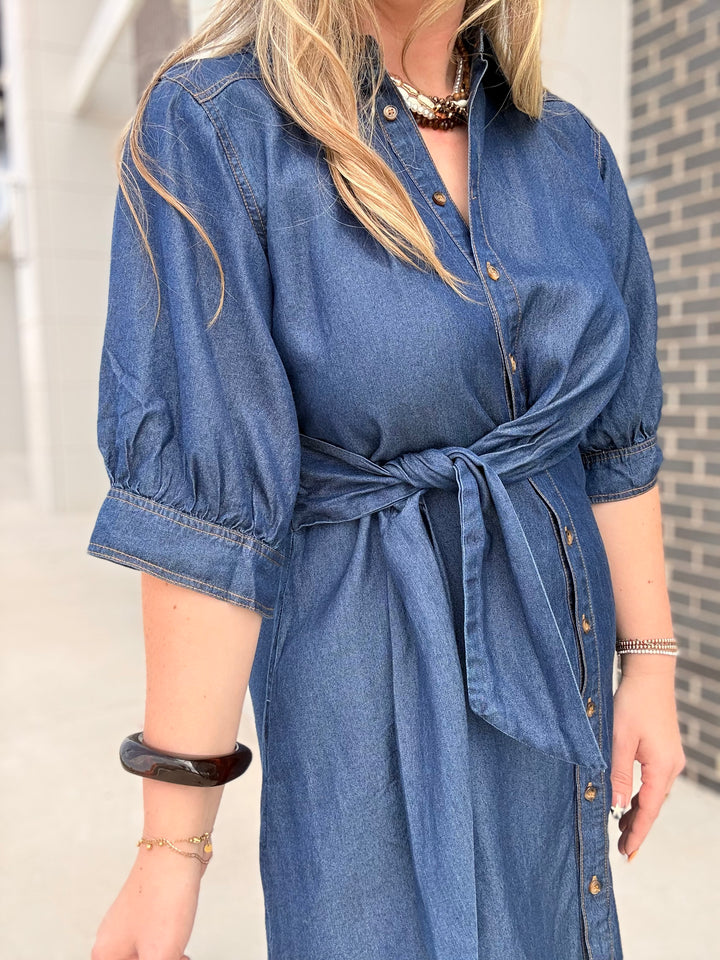 A woman wearing a blue denim shirt dress with puff sleeves, a button front, and a tie waist, photographed outdoors against a light wall and brick wall background.
