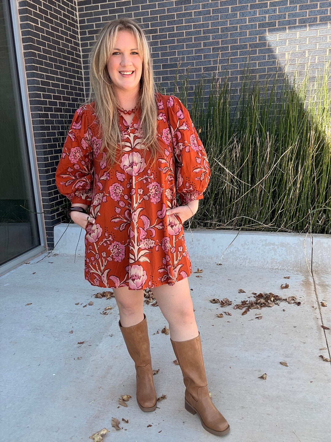 A woman wearing a rust-orange long-sleeve mini dress with a pink floral pattern and tan knee-high boots, standing outdoors in front of a dark brick wall.