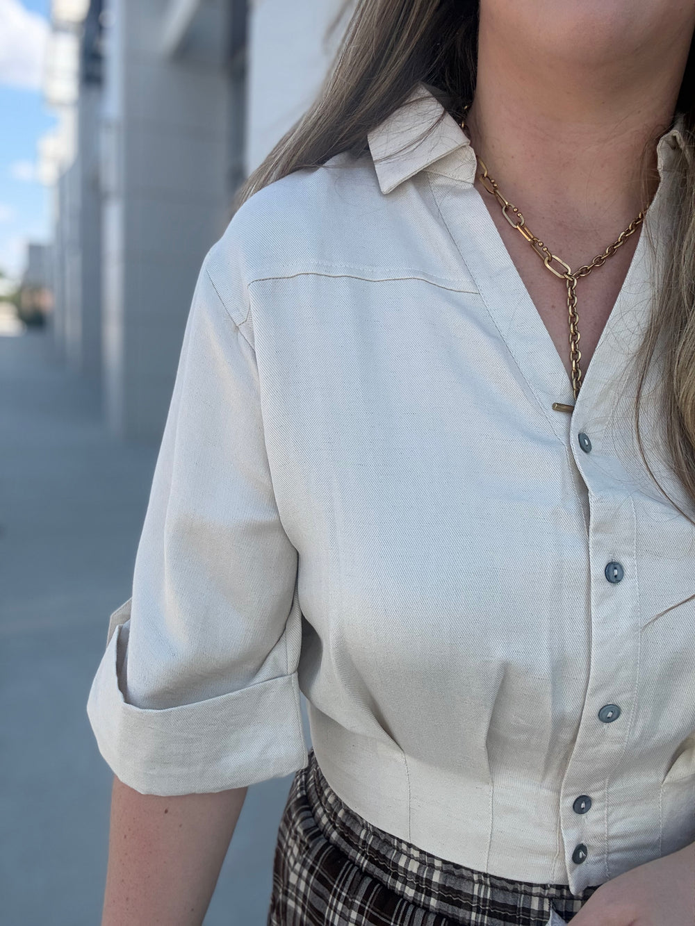 A close-up of a person wearing a light cream linen button-up shirt with rolled sleeves and a gold chain necklace, set against an outdoor blurred background.