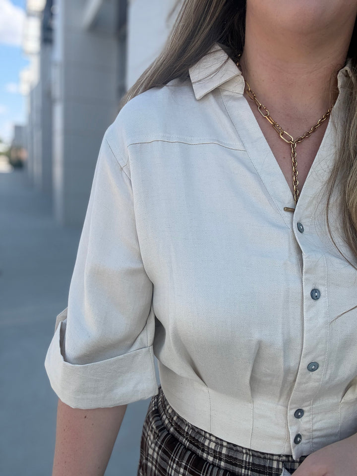 A close-up of a person wearing a light cream linen button-up shirt with rolled sleeves and a gold chain necklace, set against an outdoor blurred background.