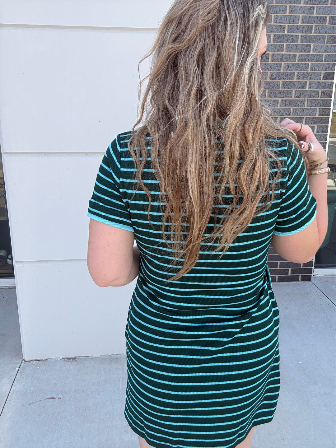 A woman viewed from behind wearing a short-sleeve dark green and teal striped t-shirt dress, standing outdoors against a white wall and brick building.