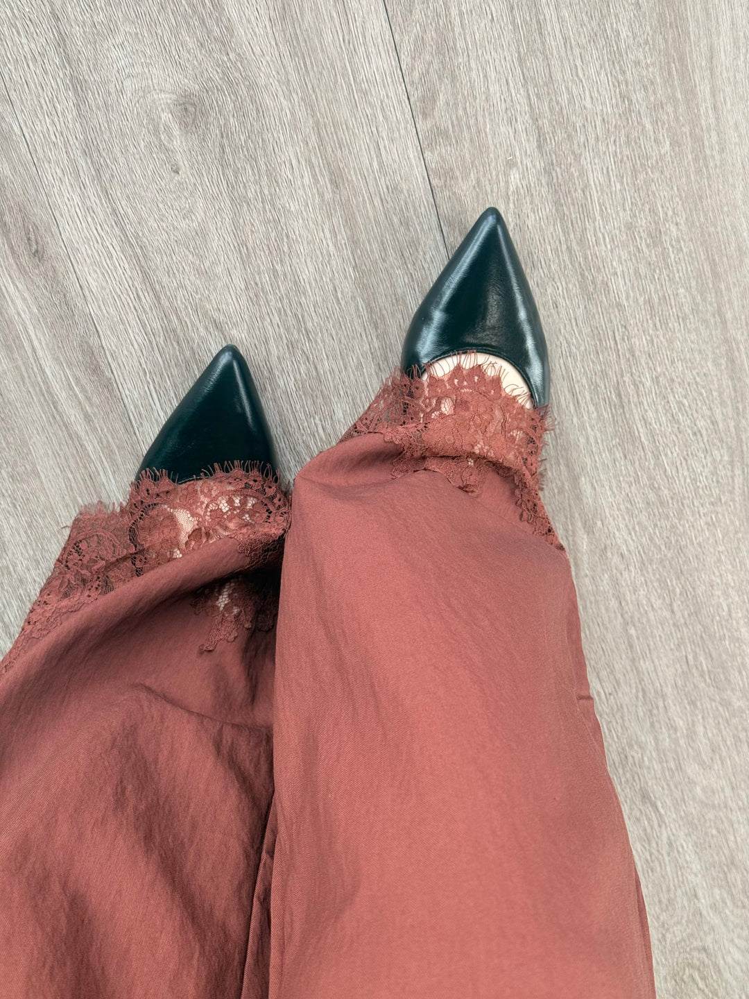 A top-down view of a person wearing dusty rose wide-leg trousers with lace trim and dark green pointed-toe flats, standing on a light gray wood-look floor.