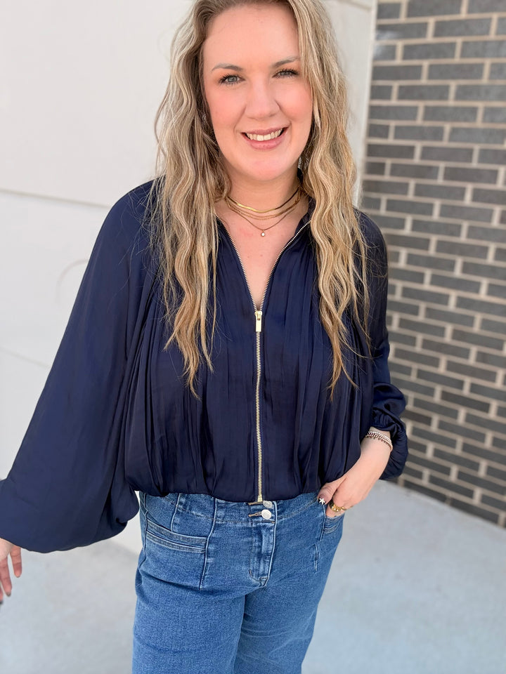 A woman with blonde hair wearing a navy blue long-sleeve blouse with a front zipper and blue jeans, standing outdoors against a white wall and a gray brick wall.