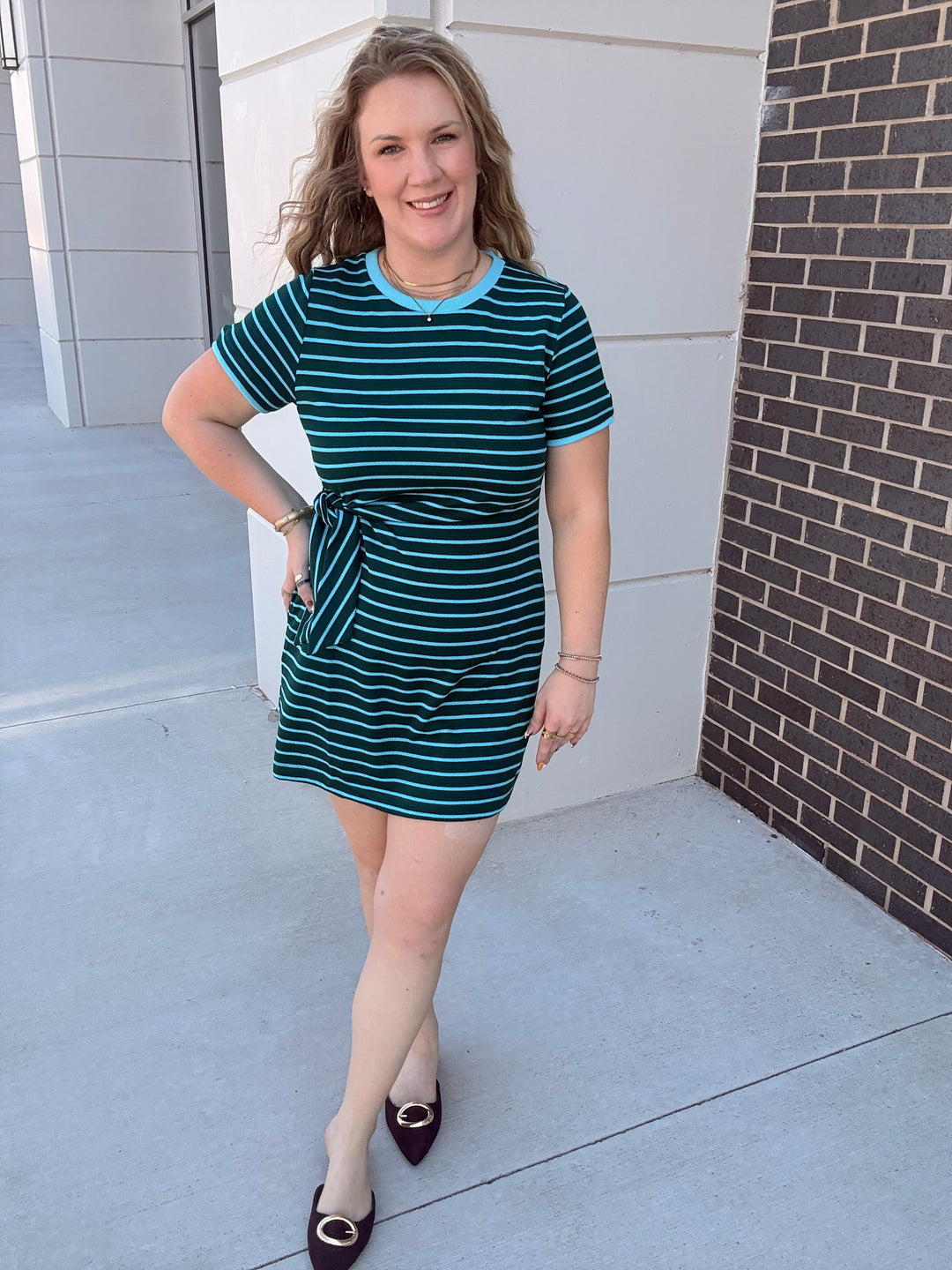 A woman modeling a short-sleeve mini dress with horizontal teal and white stripes, featuring a light blue crew neck trim and a matching side tie, paired with beige tights and dark brown flats.